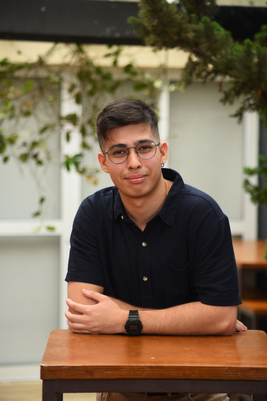 Davi Camargo, psicólogo, sorrindo, apoiado numa mesa de madeira